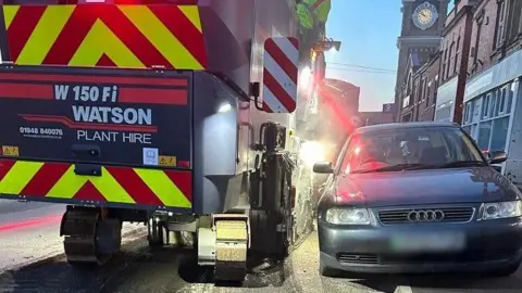 A highways resurfacing vehicle with red and yellow stripes on the back is driving around a car. The car is parked half on and half off a curb. There are buildings either side of the road. 