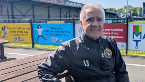 Mark Burns is sitting at a wooden picnic table a couple of meters from the edge of the pitch which is fenced off and covered in advertising boards. He's wearing a black tracksuit and has short white hair.