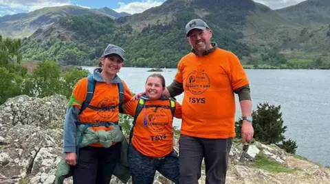 A young girl and her parents. She is in the middle, her mum and dad have their arms around her. They all have comfy hiking gear on. Behind them are huge hills and a wide lake.