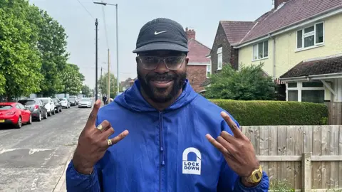 Chiedu Oraka standing in a Hull street he has a beard and wears glasses, a baseball hat and blue-hooded jacket