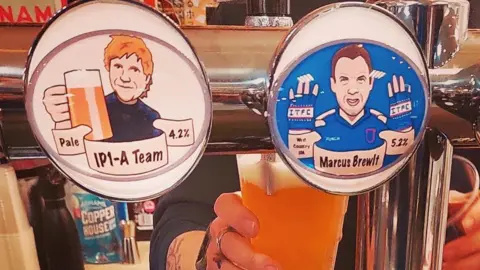 Contributed A view of several beers on draught at a pub. An Ipswich Town-themed beer and an Ed Sheeran-themed beer are on display on the front of the taps. A member of bar staff is seen behind the taps pulling a pint.