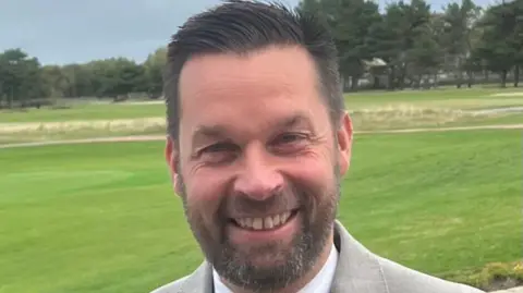 Stuart Leech Stuart Leech is photographed smiling on a golf course while wearing a grey suit jacket over a white shirt. He has greying brown hair and a trimmed beard.