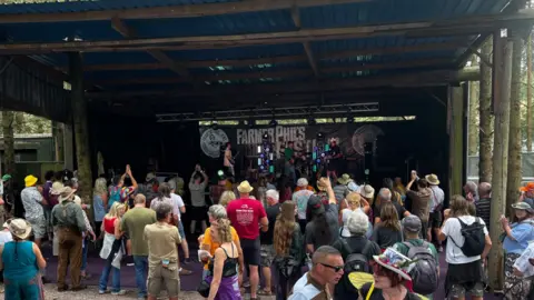 A crowd of people in colourful clothing looking towards a small stage with the words Farmer Phils on a wall at the back of it