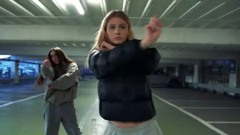 Wycombe Youth Action / Love Music Hate Violence Two people perform a choreographed dance routine in a multi-storey car park. One person stands in the foreground wearing a dark padded jacket and light-coloured tracksuit bottoms, with arms raised in a dance pose. A second person is positioned several metres behind, also mid-movement. The car park is lit by overhead fluorescent strip lights, with parking bays, support columns and a parked vehicle visible in the background.