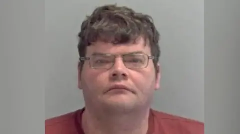 A police mugshot of Daniel Russell. He is wearing glasses and red top. 