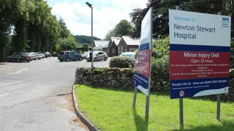 A sign at the front of Newton Stewart Hospital with a car park in the background and hospital buildings in the distance