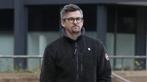 PA Media Joey Barton, who has dark grey hair styled into a quiff and wears black framed glasses and a black zip up fleece, looks into the camera with a serious expression.