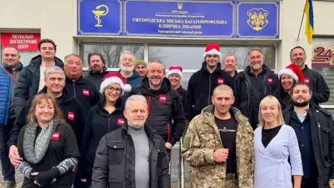 Tim Bamford About 20 people stand outside a white building with a blue and yellow sign. They are mostly wearing black coats and red Santa hats. One man is wearing camo army gear and the woman next to him is wearing a purple cardigan.