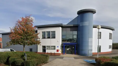 The two storey modern building is made of various blue, white and brown materials. There is a tall blue column running up the middle, and a bright blue arch over the glass doors. The building is set in from the main road, behind a pavement area with hedges and a tree on it.