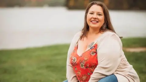 Beth Johnson squats in beige sweater and pink shirt