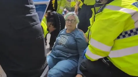 Claire Bullion A woman in blue sweatshirt and jeans sits on the ground surrounded by police officers, who appear to be arresting her.