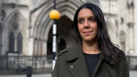 Nadine wears a dark green coat and has long black hair. She is looking off camera and standing in front of the Royal Courts of Justice. 