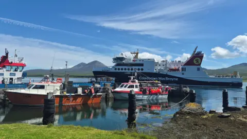 Zara MacIver Shepherd Boats in a port on the Isle of Islay. The ferry is in the water in the background and smaller fishing boats are in the foreground. 