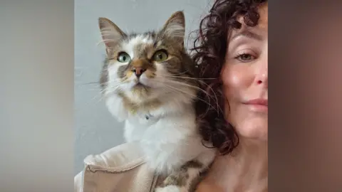 Gina holds her cat. Gina has thick curly purple hair and Sett is white with brown tortoise shell patches.