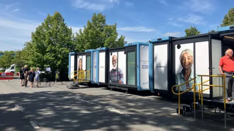 Three white demountable buildings in a sunny car park with a handful of people with grey hair stood outside.