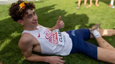 Michael Clark gives the thumbs up while laying on the grass exposing his injured foot. He is in running vest and shorts with his name on his vest bib. He is wearing sunglasses on his head. 