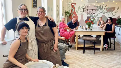 BBC Three women in black tops and brown aprons are in a pottery studio. One is sat at a potter’s wheel. There is a large table which four people and one child are sat at. There is a large mural on the back wall which includes a bottle kiln and the words “The Potter’s Nook”