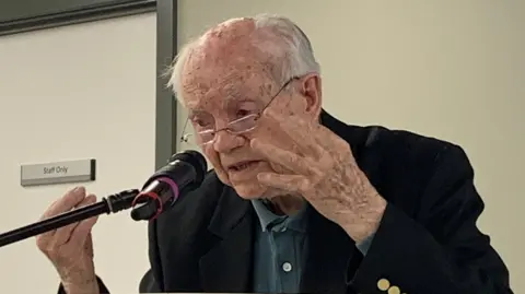 Sir Patrick Duffy wearing a turquoise polo shirt and a navy blazer is speaking animatedly into a microphone.