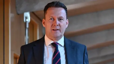 Russell Finidlay, who has short black hair, looks to his right as he walks in a wood-panelled concrete corridor. He is wearing a navy suit and tie with a white shirt. 