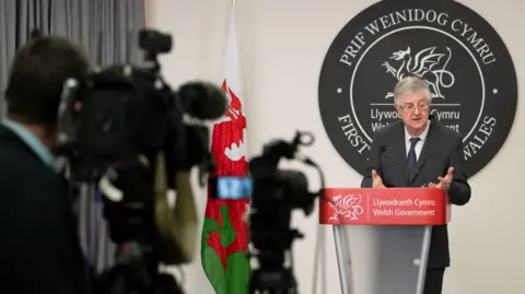 Getty Images First Minister of Wales Mark Drakeford speaks during a coronavirus press conference, standing in front of a lectern beside a Welsh national flag, and a cameraman operating a camera is seen in the foreground. 