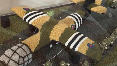 A knitted yellow, black and green camouflaged bomber with the red, white and blue RAF logo at The Longest Yarn exhibition. A number of green knitted soldiers carrying black knitted rifles are marching alongside the bomber.