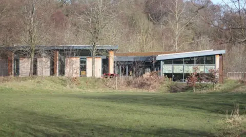 Groundworks NE & Cumbria The Land of Oak & Iron heritage centre in the Derwent Walk Country Park. The building has wood panelling and large windows. Outside is a large patch of grass where the new play park will be built.