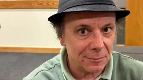 Martin Heath/BBC A person wearing a dark hat and a light-coloured collared shirt is seated indoors in a room with wood‑panelled trim, a door, and framed items on the wall.