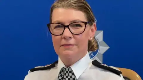 Judi Heaton, wearing a black and white check tie and a white shirt, looking serious and straight at the camera
