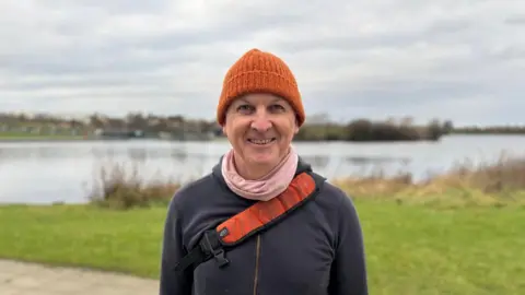 A man standing in front of a lake with an orange beanie hat on and an orange shoulder bag across his chest