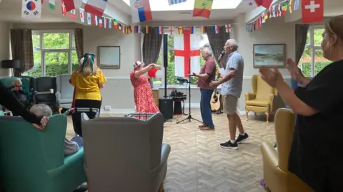 Several people are singing and dancing in the middle of a main room in a care home. There is a large England flag on the window in the back 