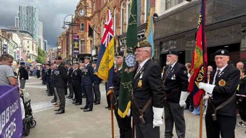A ceremonial parade taking place on Briggate in Leeds. Shopfronts, street lighting, and tall structures stretch into the background. In the foreground a line of uniformed men stand dressed in dark outfits, some wearing berets or caps, and many have medals pinned to the front of their jackets. Some are wearing white gloves and holding tall ceremonial flags upright. 