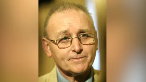 A close up image of Denis Donaldson. He has thinning, short grey hair and has a neutral expression on his face as he takes a question at a press conference. He has a pair of silver glasses on. He is wearing a tan suit jacket and a blue collared shirt.