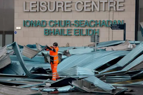 Getty Images A worker in orange reflective work clothing throwing damaged pieces of a blue/grey plastic roof outside the Helensburgh Leisure Centre