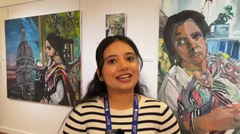 A young woman from India smiles at the camera, wearing a sweatshirt and University of Liverpool lanyard, as she stands in front of Indian paintings at the exhibition.