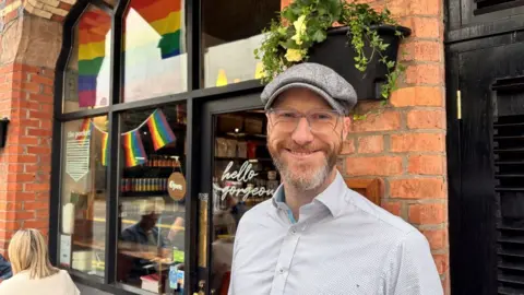 A person wearing glasses, a grey flat cap and a light patterned shirt stands outside a red brick-fronted shop. Rainbow Pride flags are displayed in the shop windows behind them, and hanging plants decorate the exterior wall above the entrance. Visible window text includes “hello gorgeous” and “open".