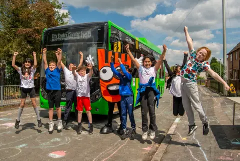 A line of children jump in the air and cheer in front of a green bus