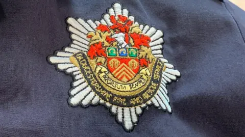 A close up of the Gloucestershire Fire and Rescue Service badge on a firefighter's navy blue shirt. The badge comprises a red and gold crest, with a black sash beneath it.