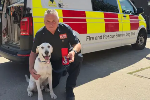 Essex County Fire & Rescue Service Graham Currie kneels down wearing a navy blue uniform, with his arm around a white dog. A fire and rescue branded van, that is white, red and yellow, is parked behind him.
