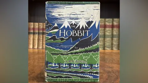 Quire Auctions A copy of The Hobbit by J.R.R. Tolkien, propped up on a brown table with a line of books behind it. The cover has illustrations of trees and mountains.