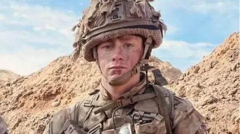 Jack in full combat gear with a jacket and camouflaged helmet in desert colours wiht steep desert mountains in the background