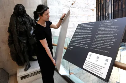 PA Media A woman dressed in black is pictured unveiling an information board. There is a large statue of a man in ceremonial robes behind her.