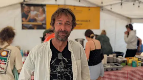 Andy Boon inside the 24 Hour Tea Tent. He is looking at the camera. People behind him are taking food and drinks form a table.