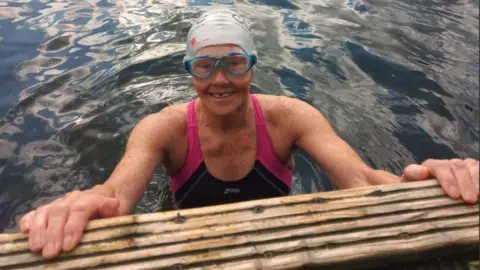 Dot Wagstaff wearing goggles and a swimming cap and swimming costume while holding onto a wooden ledge in water