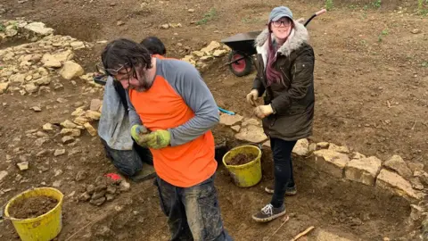 Operation Phoenix A man and a woman standing in a trench edged in stone during an excavation, quite muddy, and with yellow buckets