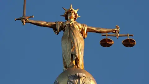 Gold Lady Justice statue holding scales of justice. The statue stands on top of a gold ball with blue sky behind. In her right hand, the statue holds a sword, the scales are in her left with a gold crown on her head.
