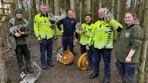 Hoo Zoo Four firefighters are smiling and have ring-tailed lemurs perched on them. There is a lemur holding onto a yellow firefighting hose on the floor. Two keepers are standing nearby smiling, one holding a lemur in his hands
