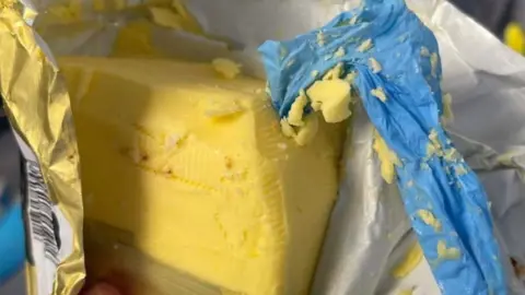 A bar of butter with a piece of blue hygienic glove poking out of it. The butter is still partially wrapped in its gold foil wrapping.