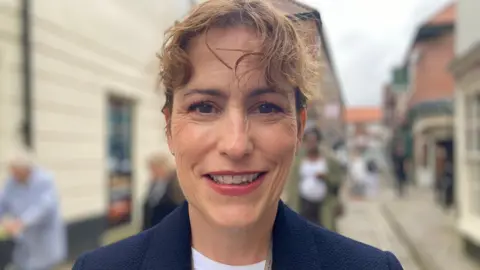 Victoria Atkins is wearing a navy jacket, copper-coloured lipstick and her hair is tied back. She is standing in a town centre smiling into the camera but the background is out of focus and blurred.