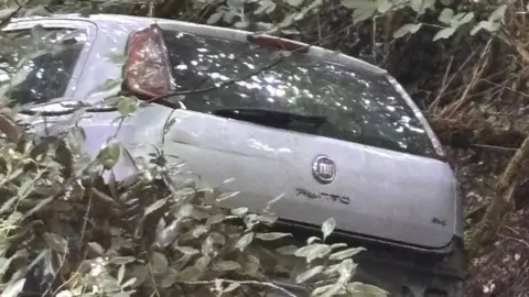 St Austell Cricket Club A silver Fiat Punto car left damaged and abandoned in a woodland area. The car is missing its rear bumper and number plate and is amongst trees and branches. 