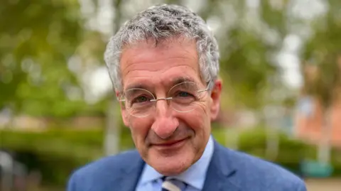 Martin Giles/BBC A head and shoulders image of a man wearing a blue suit and a blue shirt and glasses. He is smiling and looking into the camera.